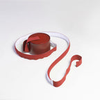 A red webbing strip winds out from a round red base on a white surface, with a white backing visible beneath on this Clérachic floor lamp.