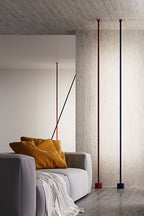 A red and a blue Clérachic floor lamp rise to the ceiling in a concrete room, with a grey sofa and ochre cushions in front.