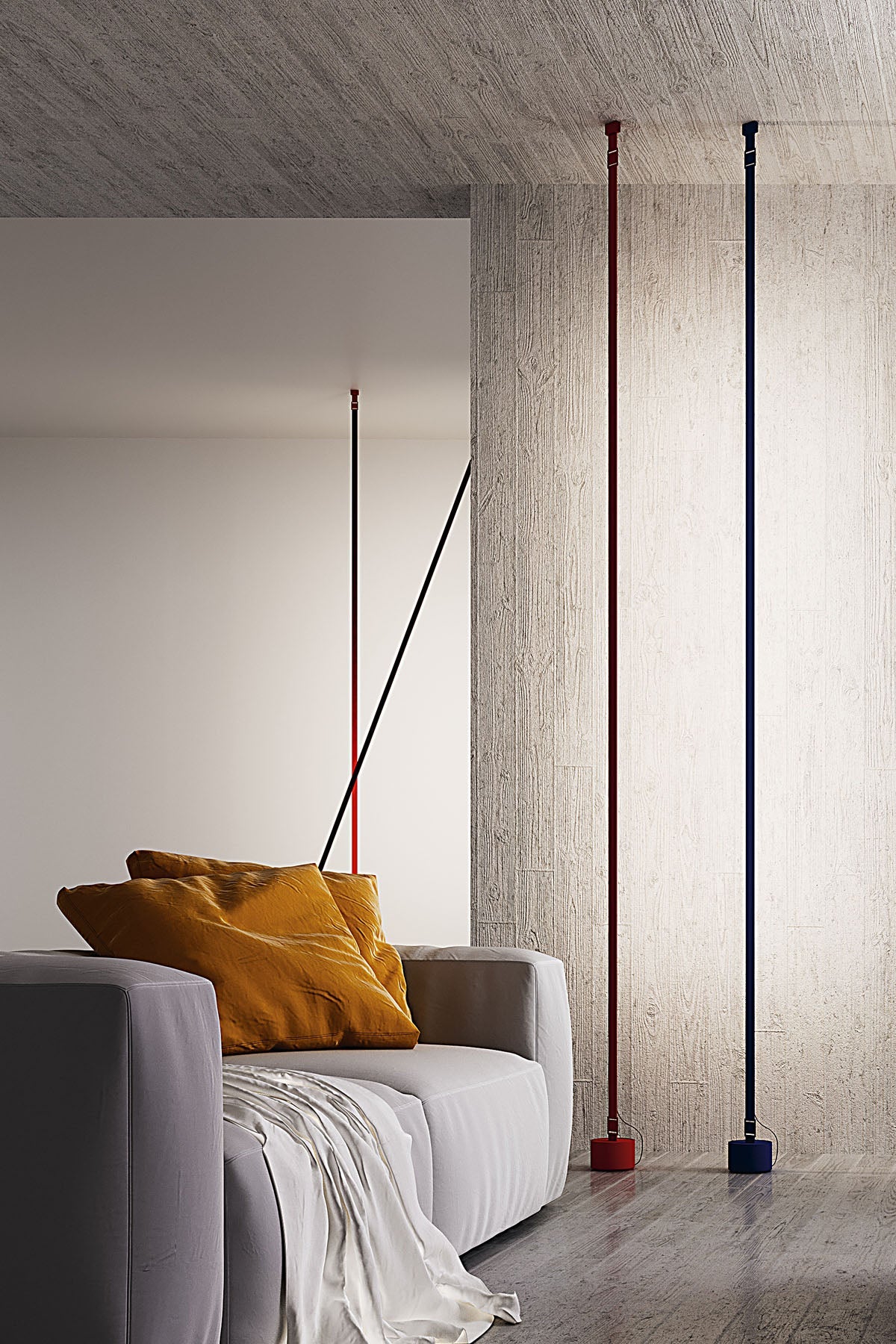 A red and a blue Clérachic floor lamp rise to the ceiling in a concrete room, with a grey sofa and ochre cushions in front.