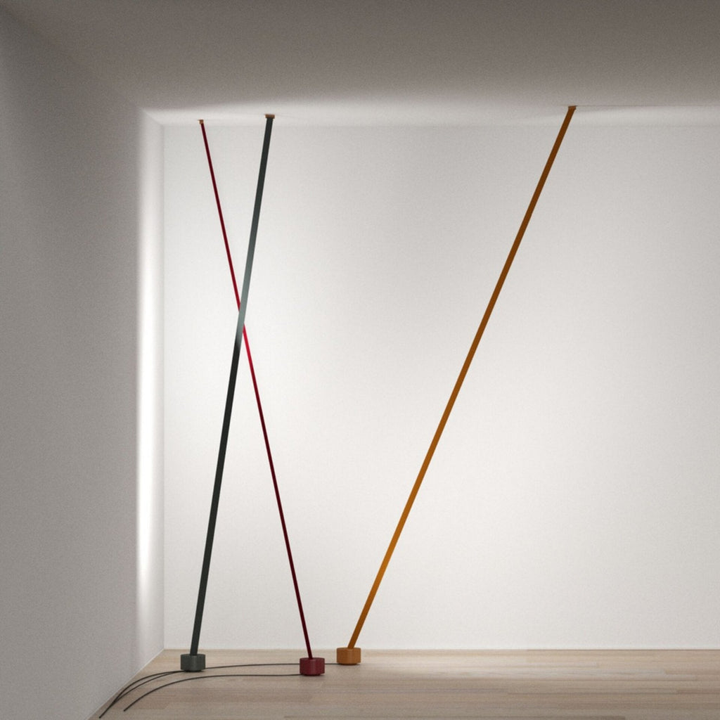 Three Clérachic floor lamp strips in grey, red and ochre lean from the ceiling in a white room, their cords running across a wood floor.