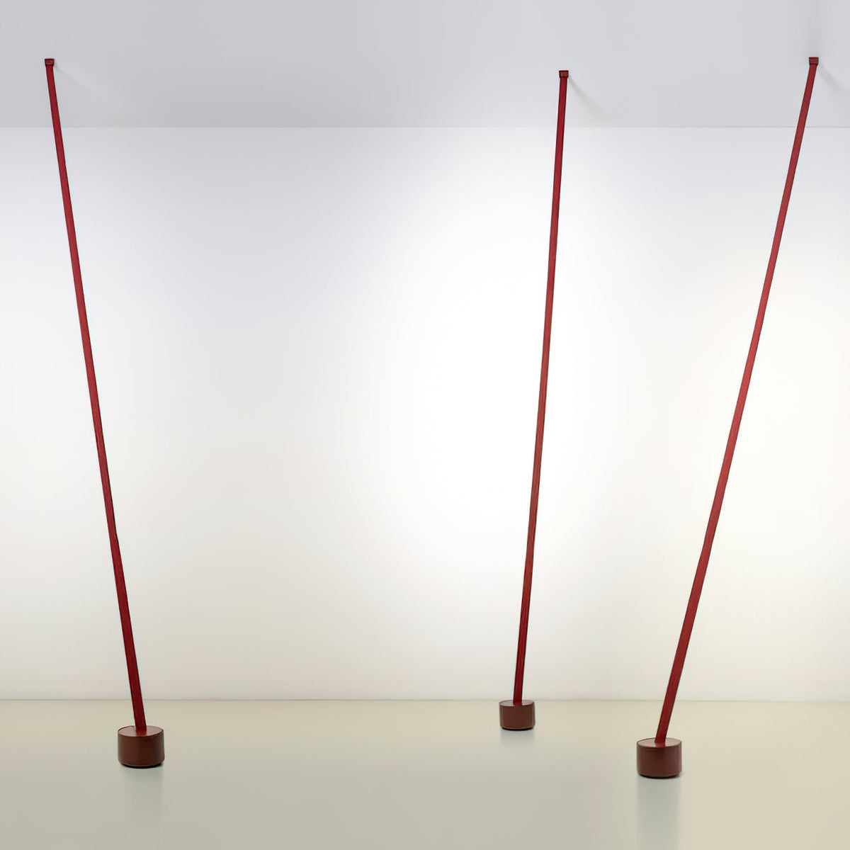 Three red Clérachic floor lamp strips stand at slight angles in a bright white room, each on a dark round base.