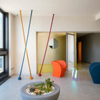 In a bright lobby with an orange armchair and a planter, three Clérachic floor lamp strips in ochre, blue and red lean at angles.