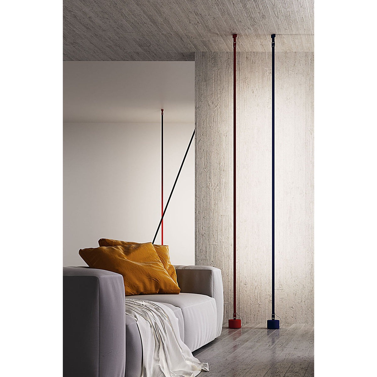 A red and a blue Clérachic floor lamp rise upright beside a concrete pillar, with a grey sofa and ochre cushions in front.