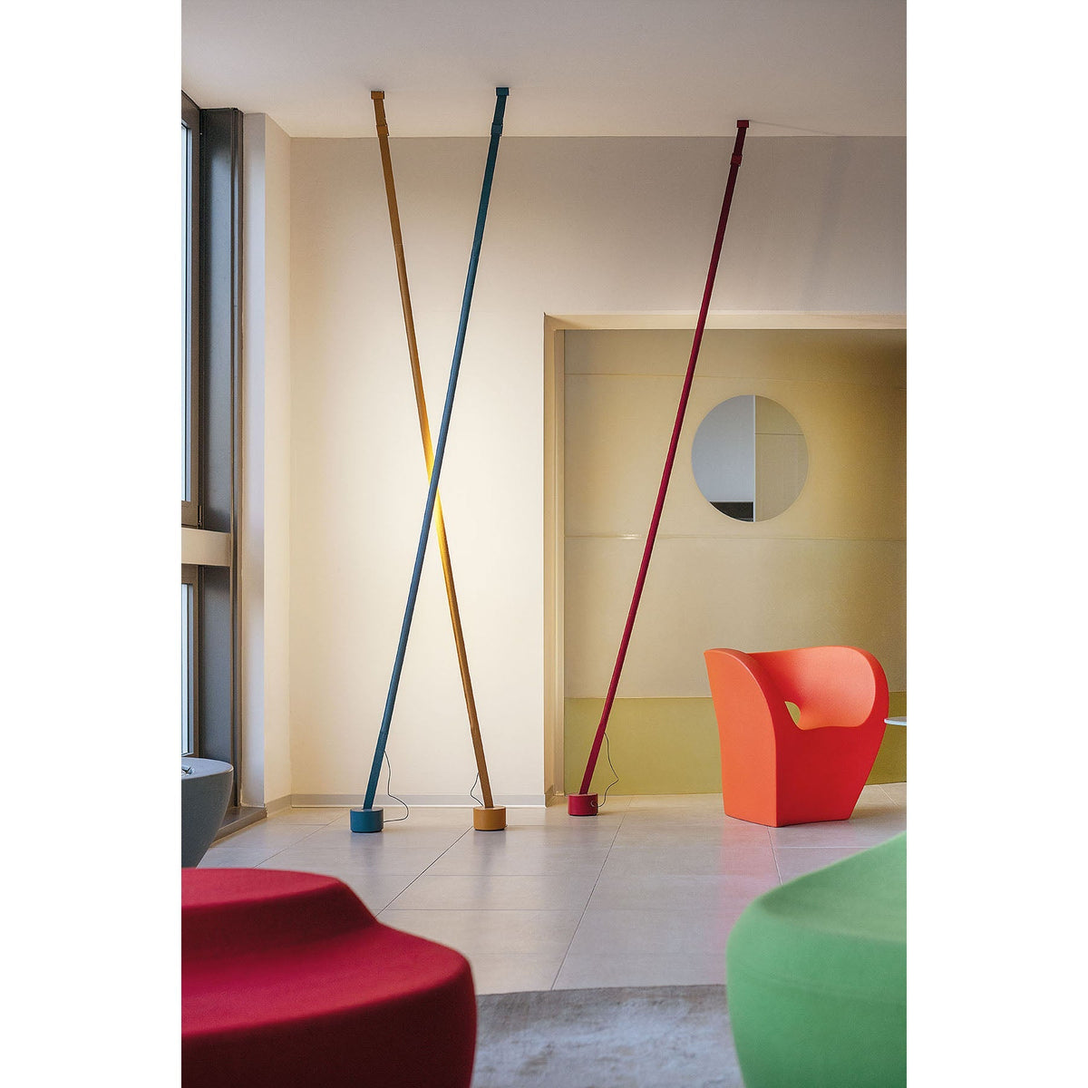 Ochre, blue and red Clérachic floor lamp strips lean in a bright lobby with an orange armchair and red and green seats.