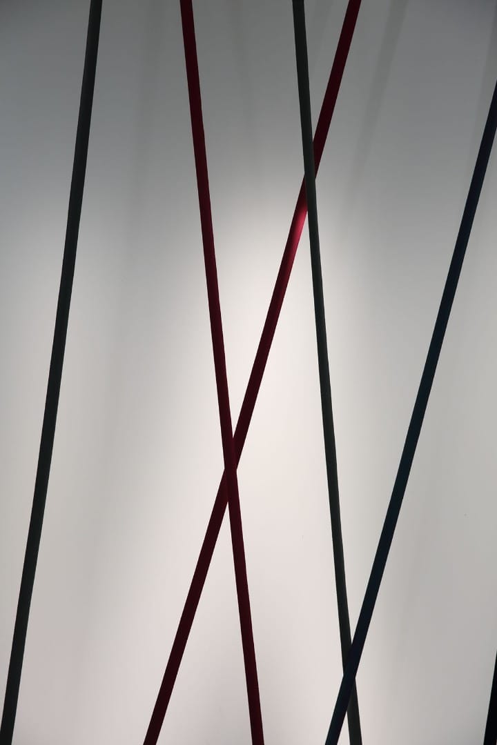 Black, red and dark grey Clérachic floor lamp strips cross at different angles in front of a softly lit grey wall.