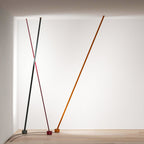 A grey, a red and an orange-brown Clérachic floor lamp strip lean in a white corner, with their cords across a pale wood floor.