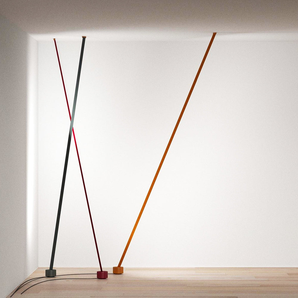 A grey, a red and an orange-brown Clérachic floor lamp strip lean in a white corner, with their cords across a pale wood floor.