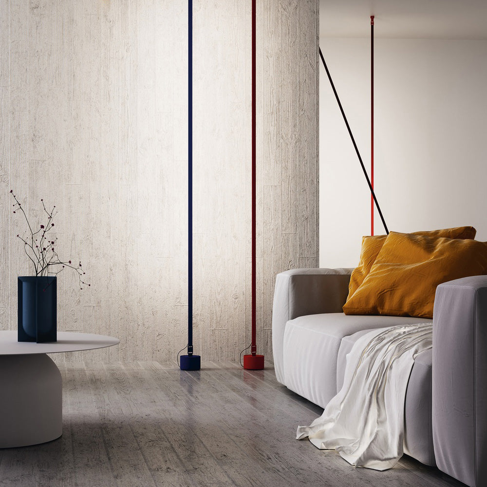 A blue and a red Clérachic floor lamp stand straight beside a grey sofa with ochre cushions, against a concrete wall.