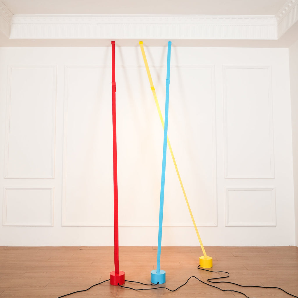 A red, a light blue and a yellow Clérachic floor lamp strip lean against a white panelled wall, their black cords across a wood floor.