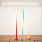 A red, a light blue and a yellow Clérachic floor lamp strip lean against a white panelled wall, their black cords across a wood floor.
