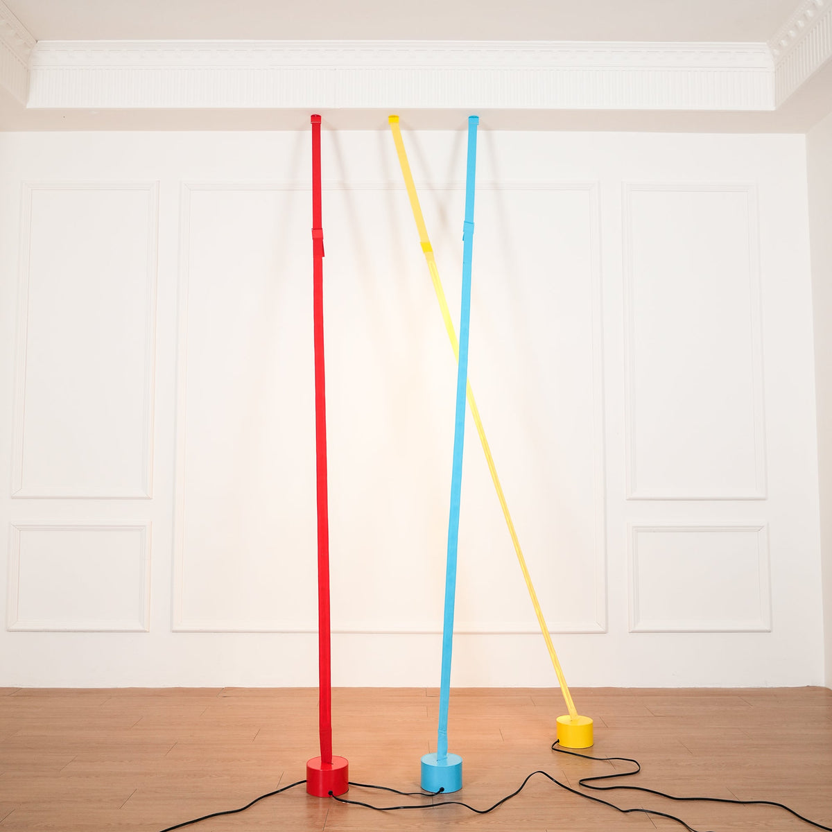 A red, a light blue and a yellow Clérachic floor lamp strip lean against a white panelled wall, their black cords across a wood floor.