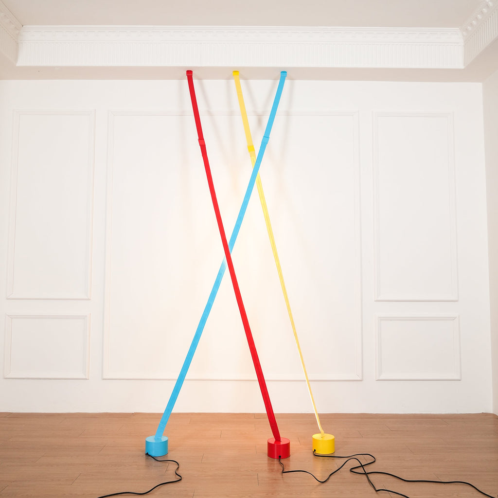 Red, light blue and yellow Clérachic floor lamp strips cross at the ceiling edge of a white panelled room, with a warm glow behind.