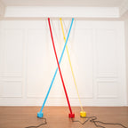 Red, light blue and yellow Clérachic floor lamp strips cross at the ceiling edge of a white panelled room, with a warm glow behind.