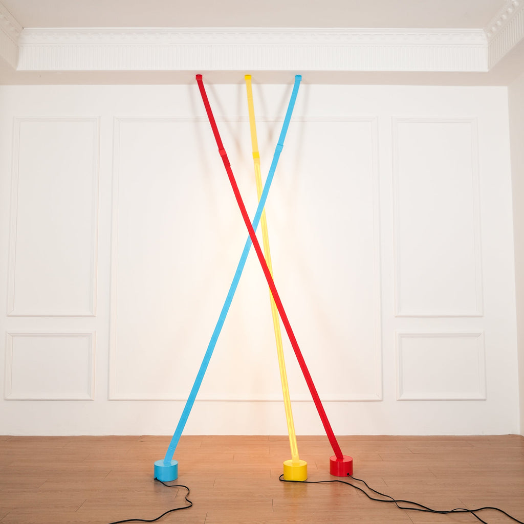 Light blue, yellow and red Clérachic floor lamp strips lean from the ceiling moulding of a white room, glowing warm on the wall.