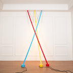Light blue, yellow and red Clérachic floor lamp strips lean from the ceiling moulding of a white room, glowing warm on the wall.