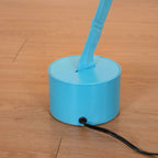 Close view of a Clérachic light blue round base on a wood floor, with the webbing strip rising from a slot and a black cord leaving the side.