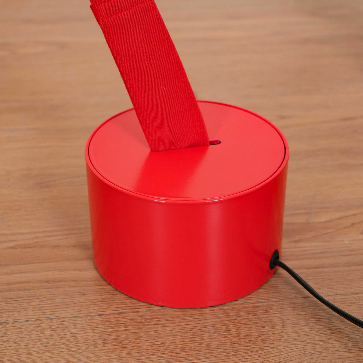 Close view of a red round base on a wood floor, with the Clérachic floor lamp strip rising from a slot and a black cord at the side.
