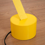 Close view of a yellow round base on a wood floor, with the Clérachic floor lamp strip rising from its slot and a black cord beside it.