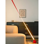 A red Clérachic floor lamp strip leans across a living room, lit with a warm bright line above a beige sofa and a black sideboard.