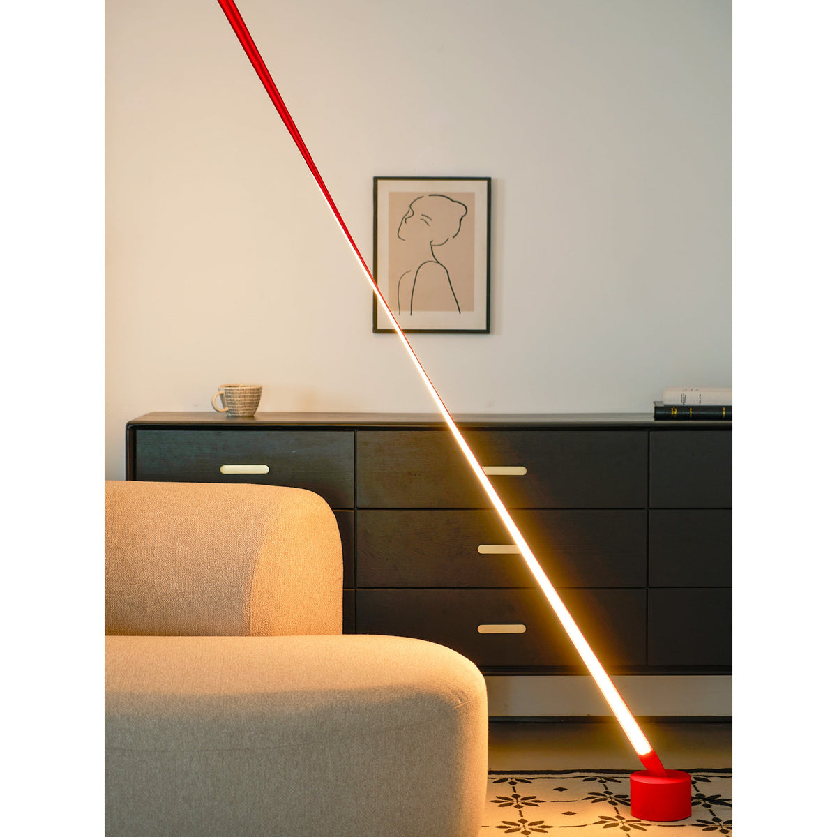 A red Clérachic floor lamp strip leans across a living room, lit with a warm bright line above a beige sofa and a black sideboard.