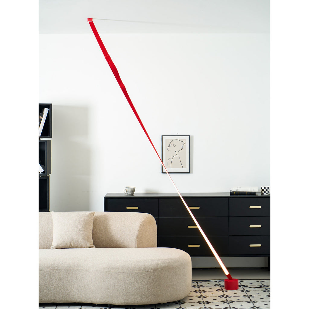 A red Clérachic floor lamp strip stretches from the ceiling to a red base on a patterned floor, beside a curved beige sofa.