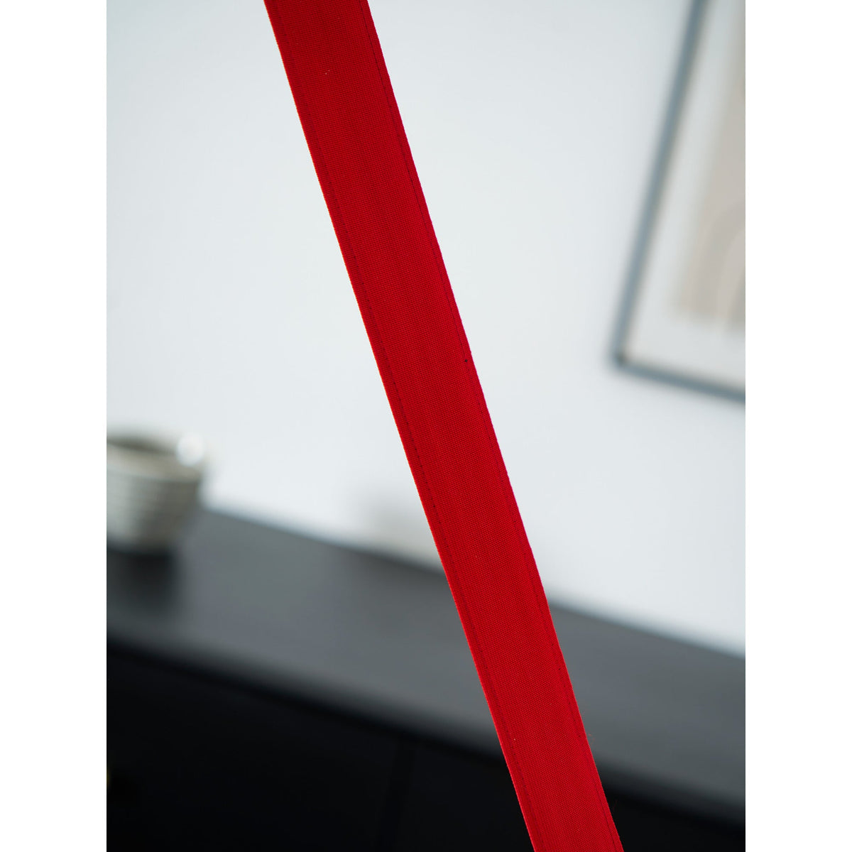 Close view of the red webbing of this Clérachic floor lamp, with a blurred black sideboard and picture frame behind.