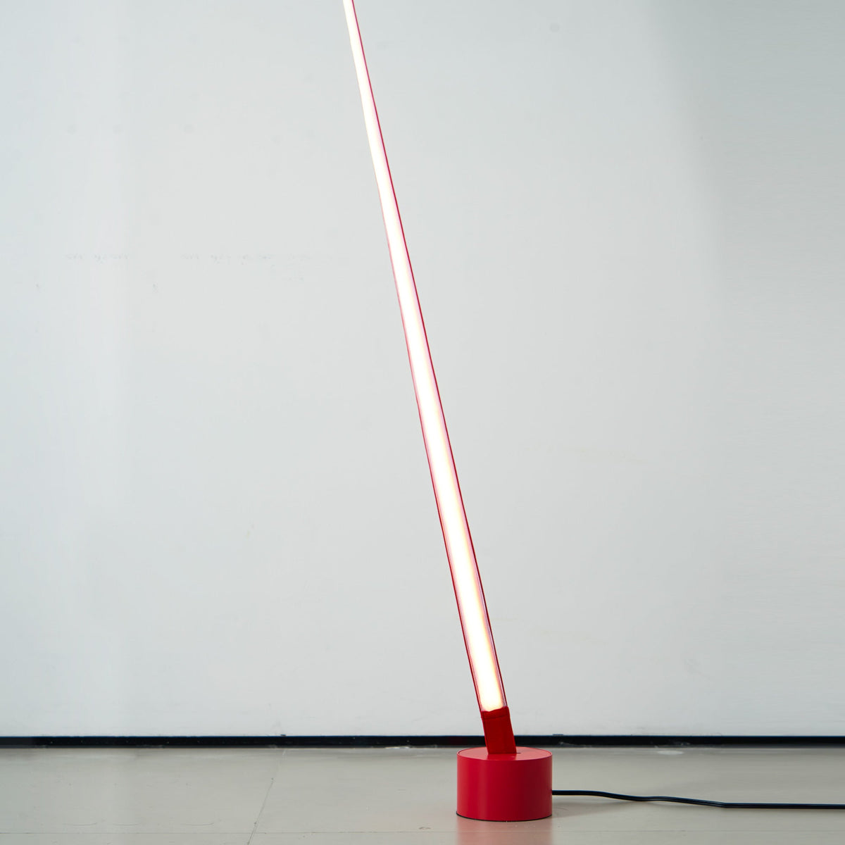 The lit lower end of a red Clérachic floor lamp strip rises from a round red base on a pale floor, glowing white along its length.