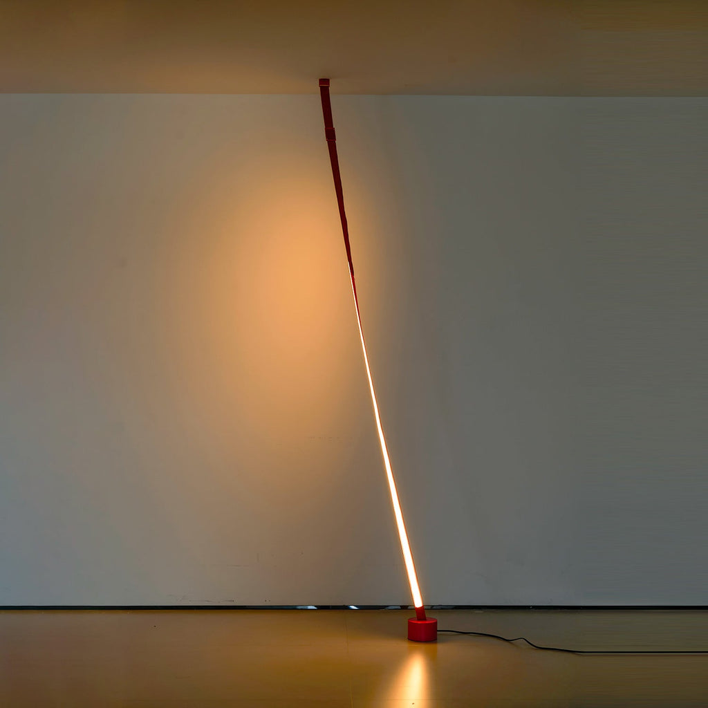 A red Clérachic floor lamp leans from the ceiling to a round base in a dim gallery room, lit with a warm line and a glow on the wall.