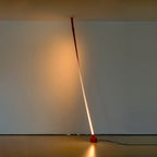 A red Clérachic floor lamp leans from the ceiling to a round base in a dim gallery room, lit with a warm line and a glow on the wall.