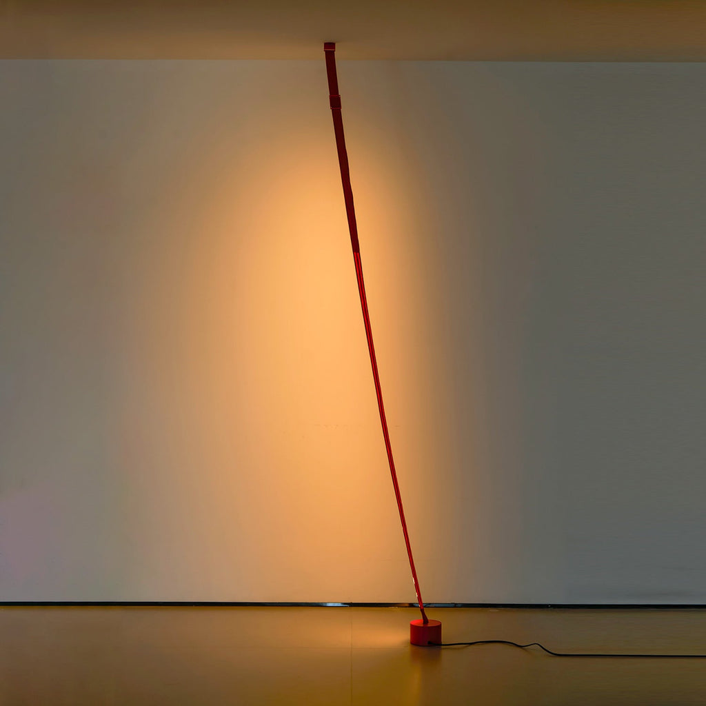 An unlit red Clérachic floor lamp strip curves gently from the ceiling to a round base in a dim room with a warm glow behind.