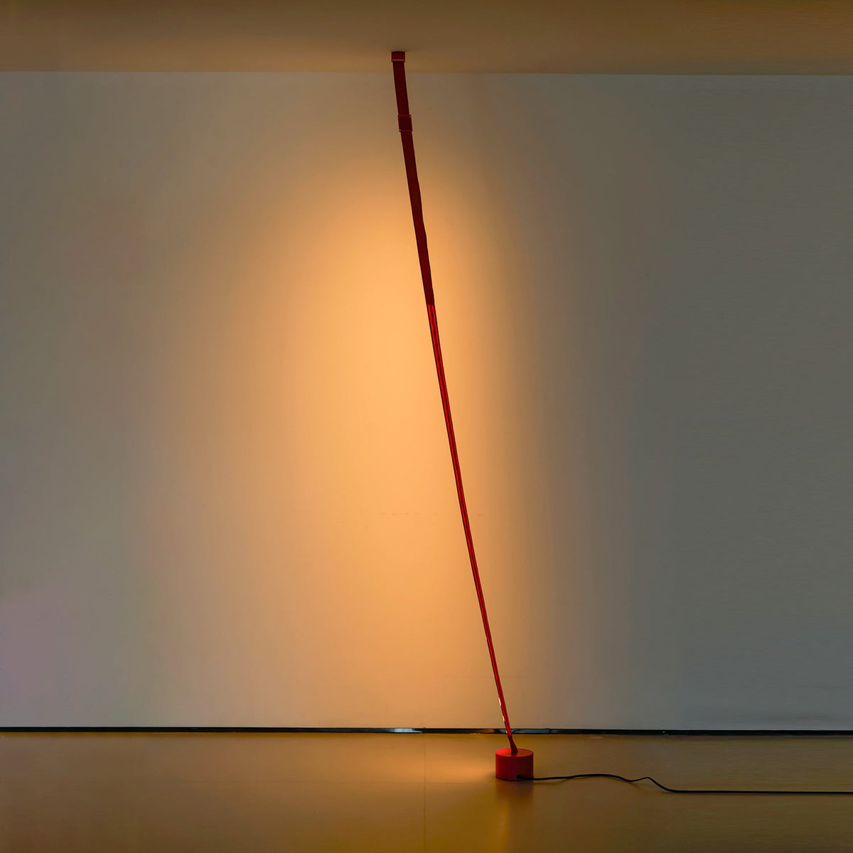 An unlit red Clérachic floor lamp strip curves gently from the ceiling to a round base in a dim room with a warm glow behind.
