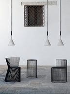 Three small grey Clérachic cone pendants at different heights in front of a white wall, above three black wire stools.