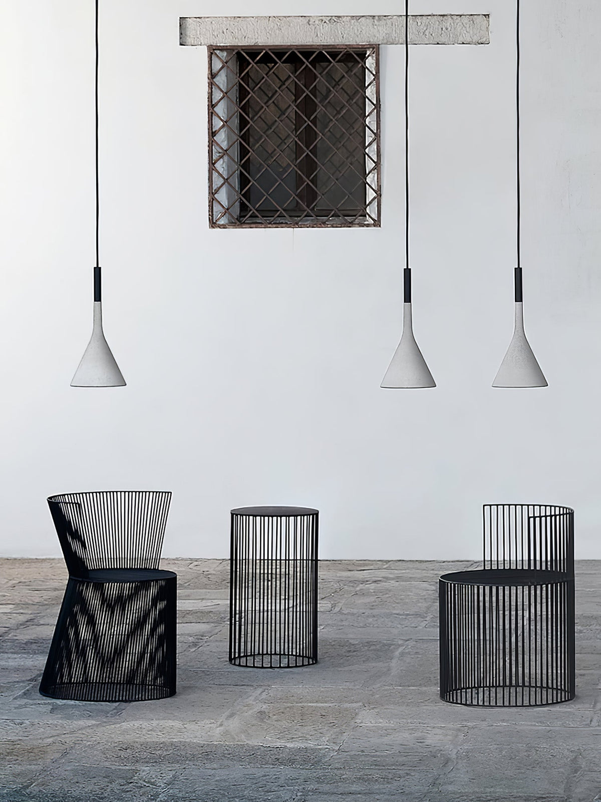 Three small grey Clérachic cone pendants at different heights in front of a white wall, above three black wire stools.