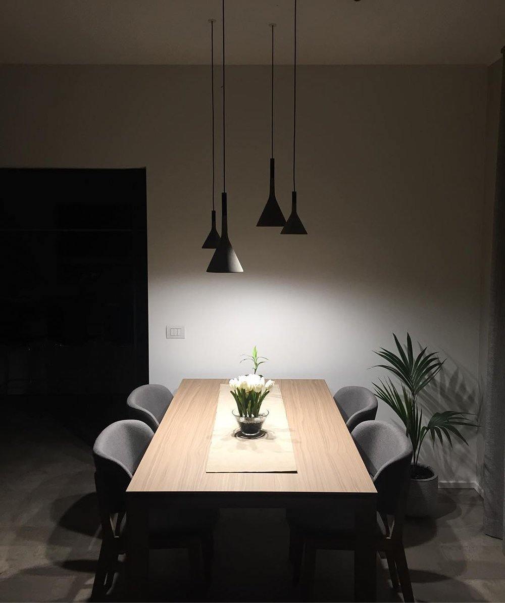 Three black Clérachic cone pendants lit over a long wooden table with grey chairs, in a dim room with a plant at the side.
