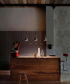 Three grey Clérachic cone pendants lit over a rustic wooden counter, against a brick-tiled wall in a warm evening light.