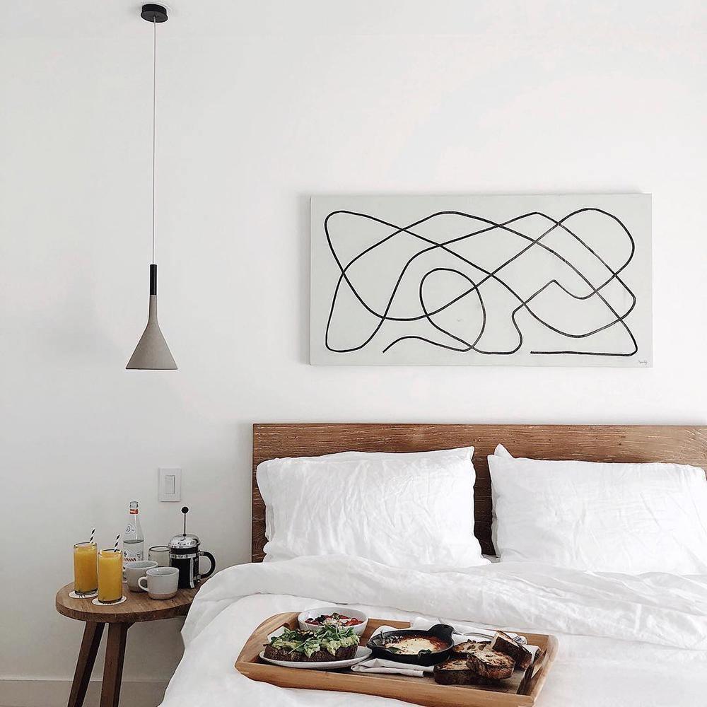 A single grey Clérachic cone pendant hung over a bed with white linen, beside a line-drawing canvas on a white wall.