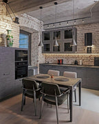 Three Clérachic cone pendants over a table in a loft kitchen with exposed brick walls, a track of spotlights and grey chairs.