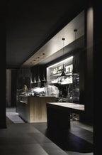 A row of small Clérachic cone pendants in a dark bar and kitchen, lit above a wooden counter with shelves of bottles behind.