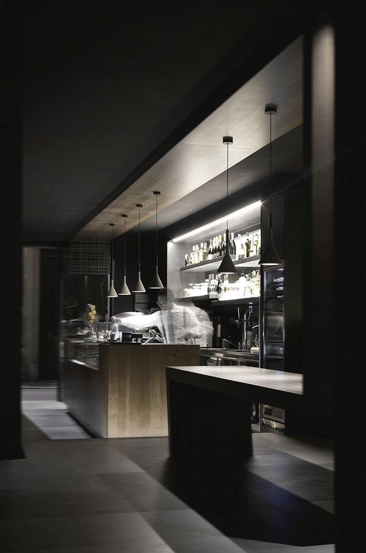 A row of small Clérachic cone pendants in a dark bar and kitchen, lit above a wooden counter with shelves of bottles behind.