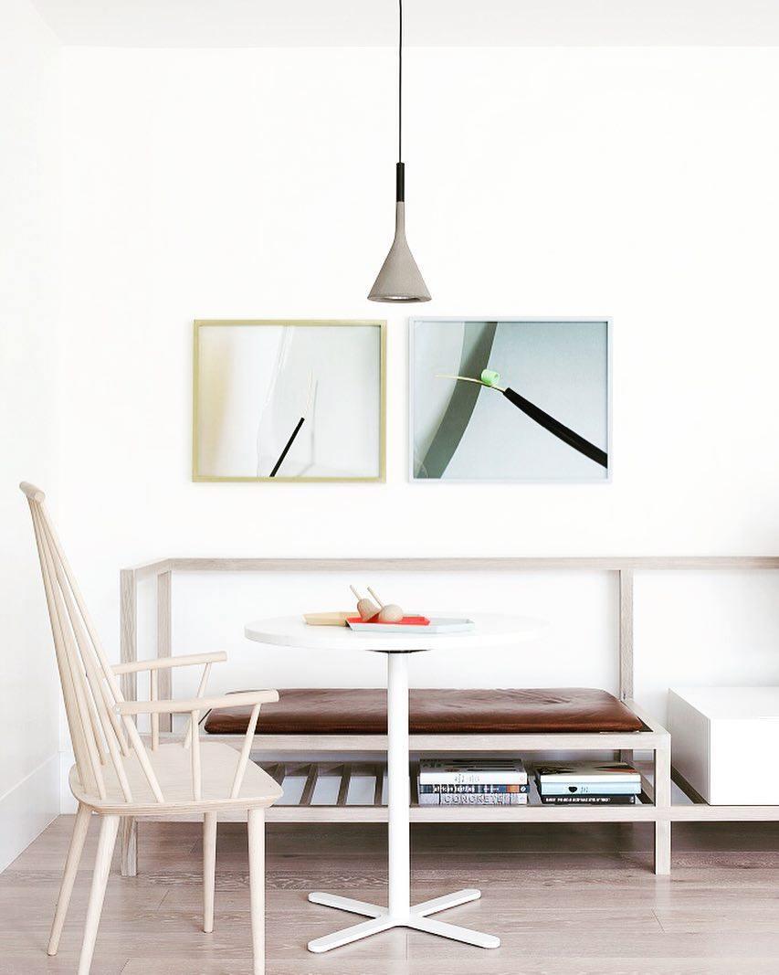 A single grey Clérachic cone pendant over a round white pedestal table, with two framed prints on a white wall and a bench seat.