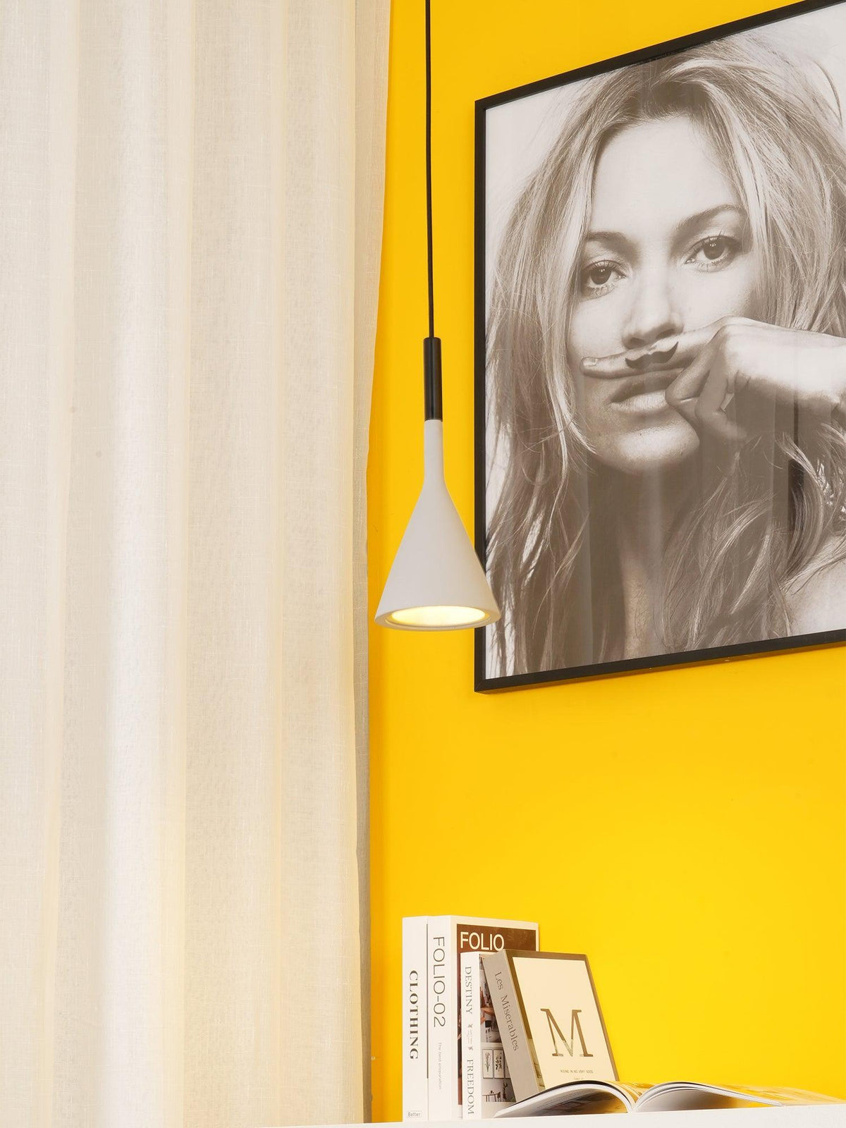 A single pale Clérachic cone pendant with a black cord, shown against a yellow wall with a framed portrait and a stack of books below.