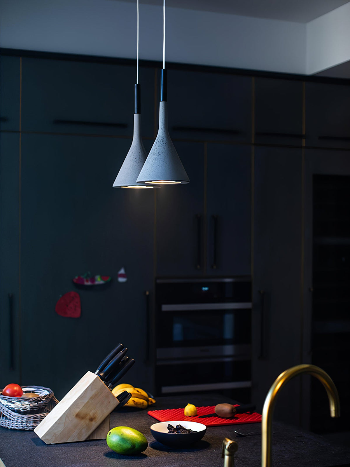 Two pale grey Clérachic cone pendants hung low in a dark kitchen with black cabinets and a counter with fruit and a knife block.