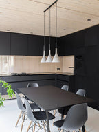 Three white Clérachic cone pendants on a long black bar above a black kitchen table, with grey chairs and a light wood ceiling.