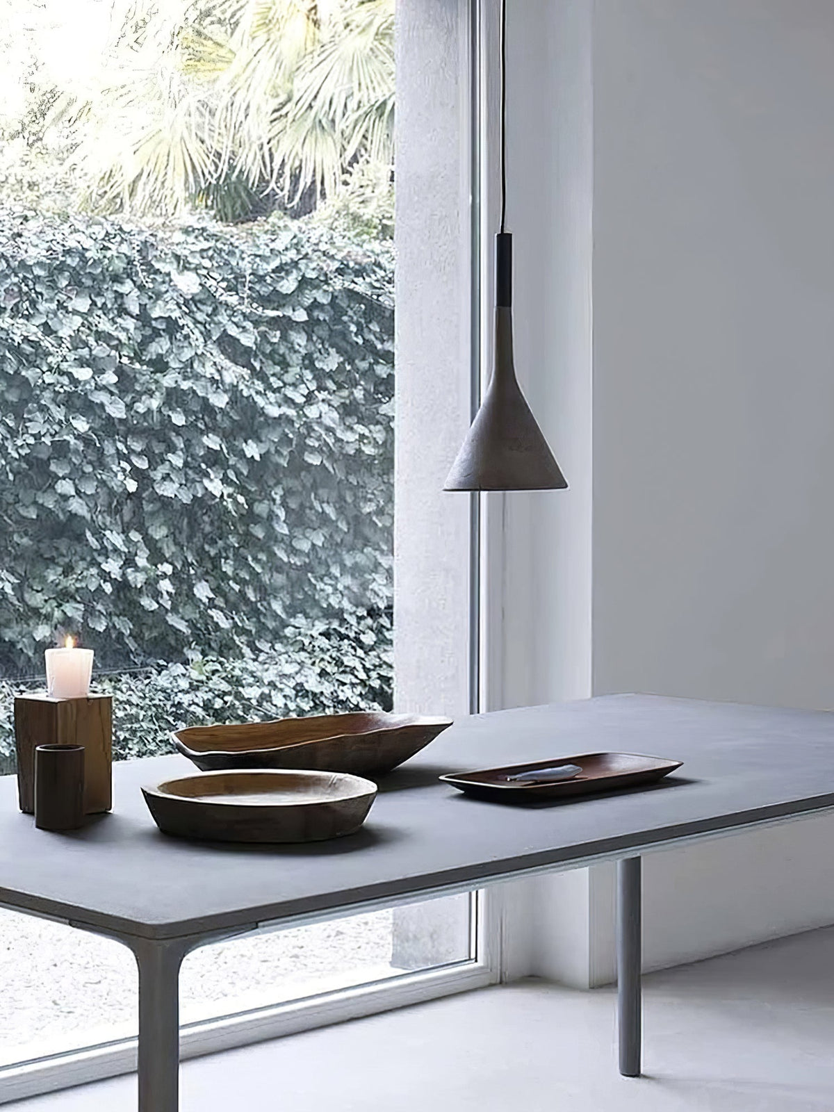 A dark grey Clérachic cone pendant hung above a stone-grey table with wooden bowls and a candle, in front of a glass wall and ivy.