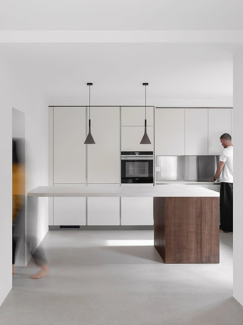Two Clérachic cone pendants lit above a pale island in a white kitchen, with tall cabinets and a person standing at the counter.