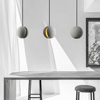 A cement-grey Clérachic sphere pendant with a lit crescent slot hangs between two plain ones above a long table and stool.