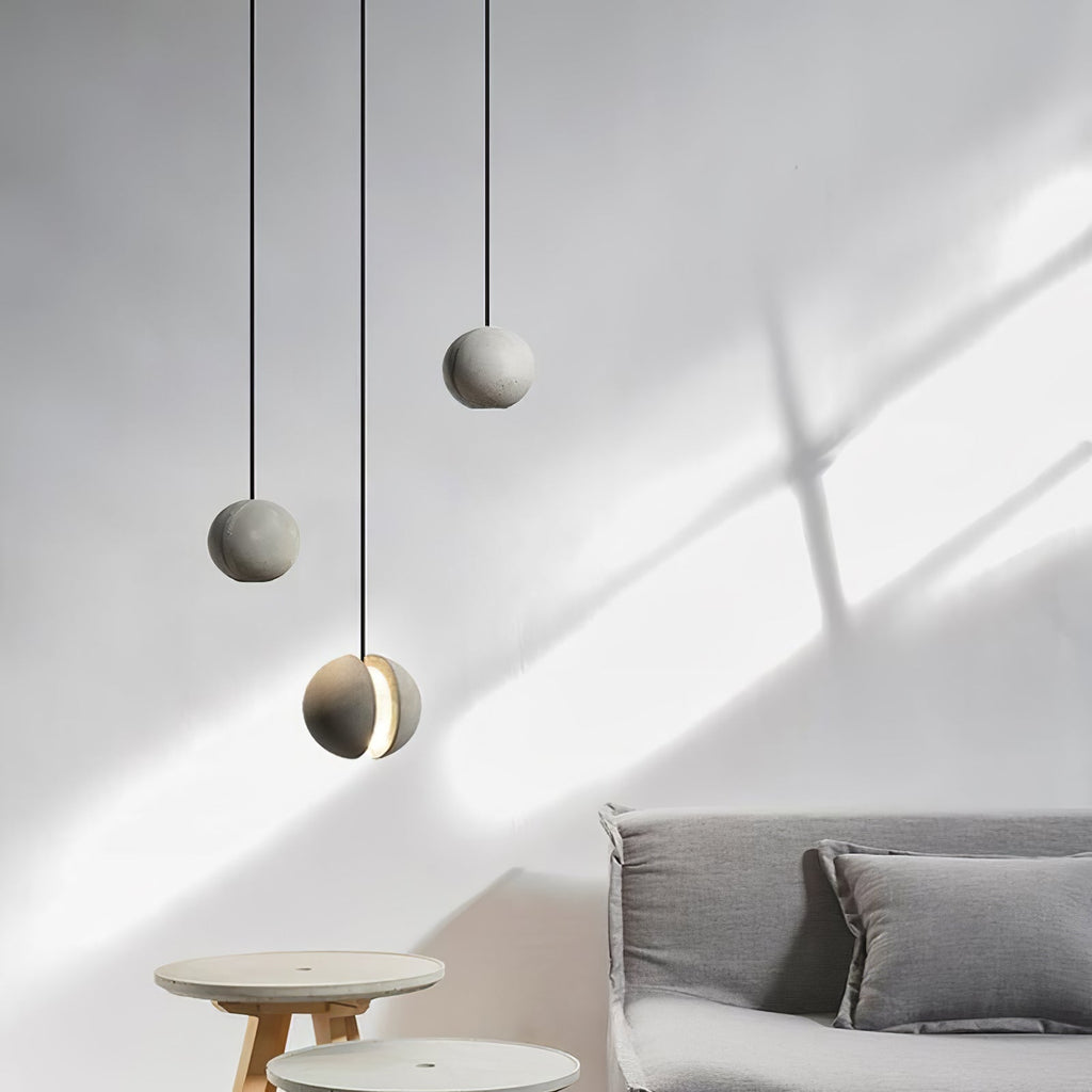 Soft shadows cross a pale wall behind three small Clérachic concrete pendants, one glowing, above stools and a grey sofa.