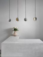 A row of four cement-grey Clérachic spheres hangs over a pale plinth topped with a patterned vase of white flowers.