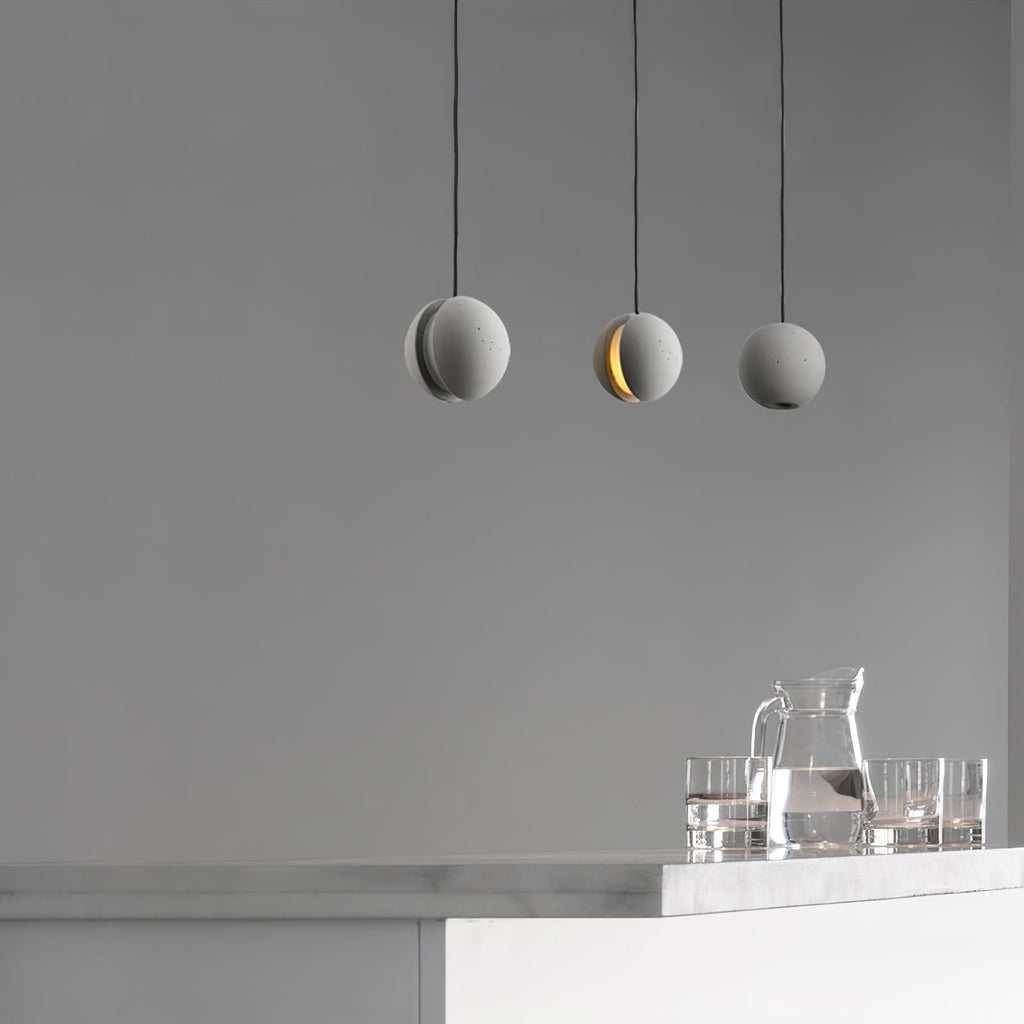 Three Clérachic concrete pendants hang against a grey wall, the middle one lit, above a counter with a glass jug and tumblers.