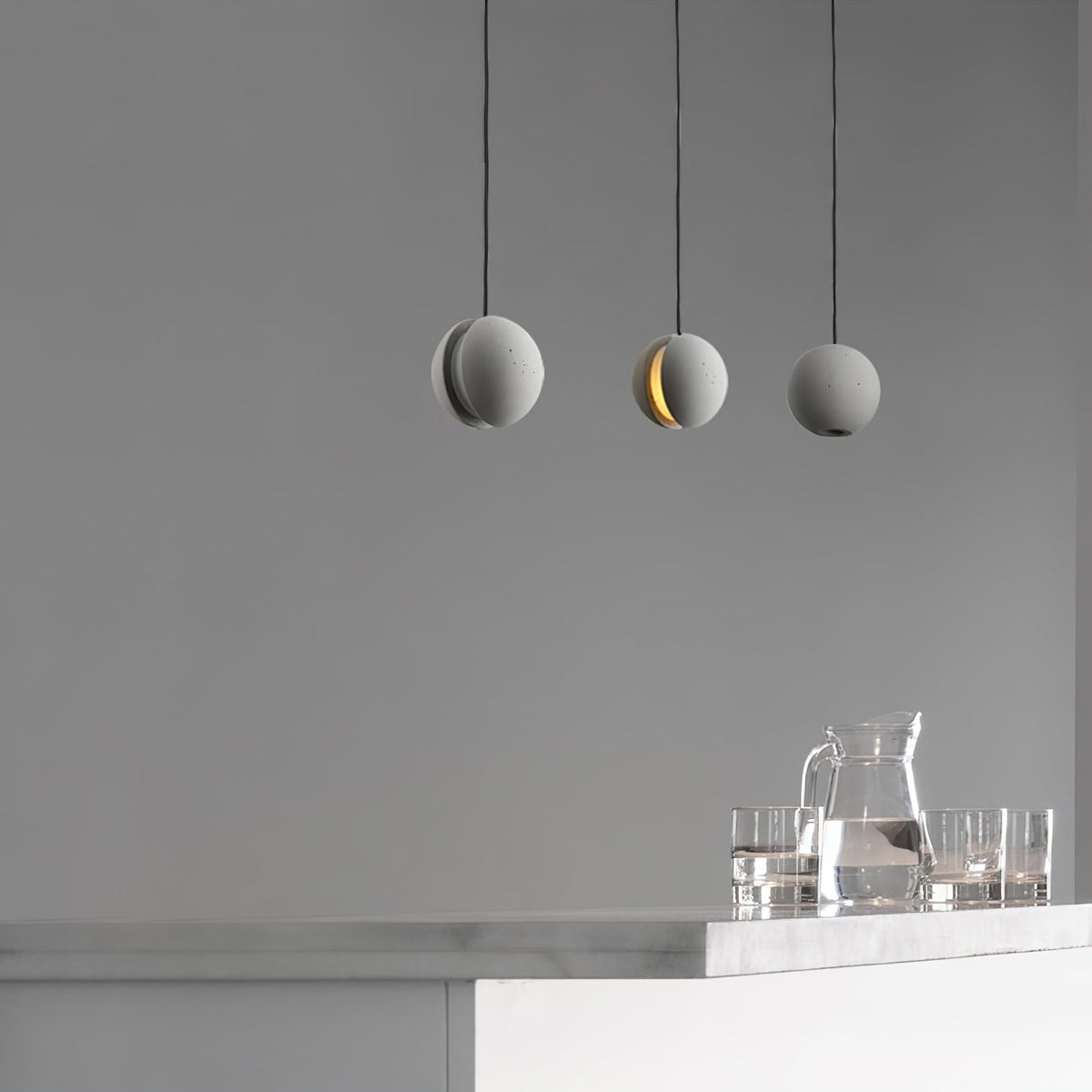 Three Clérachic concrete pendants hang against a grey wall, the middle one lit, above a counter with a glass jug and tumblers.